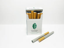 Load image into Gallery viewer, Product displayed resembles a white classic cigarette pack with a large seed/stem logo that is various shades of textured green.  The company logo is on the front flap and the text is in green label with font Manrope.  Front of pack is shown open with hemp smokes visible.  Two smokes lay in the foreground showing either end of the smokes to show the filter and the flower inside. 