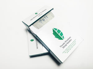 Product displayed resembles a white classic cigarette pack with a large seed/stem logo that is various shades of textured green.  The company logo is on the front flap and the text is in green label with font Manrope.  Pack is shown in plan view, front flap is partially open to reveal the silver foil that is labeled 'Pull' There are two packs shown on angled on top of the other.  