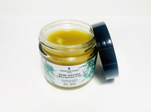 open glass jar reaveals golden coconut oil infused with hemp with white label includes green botanical drawings of hemp or cannabis plants with label text in black along with company logo.  Jar lid is black.  Photo is on a white background.